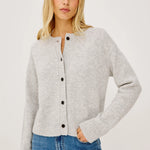 A woman wearing a light gray heathered knit cardigan with black buttons and blue jeans, photographed against a clean off-white studio background.