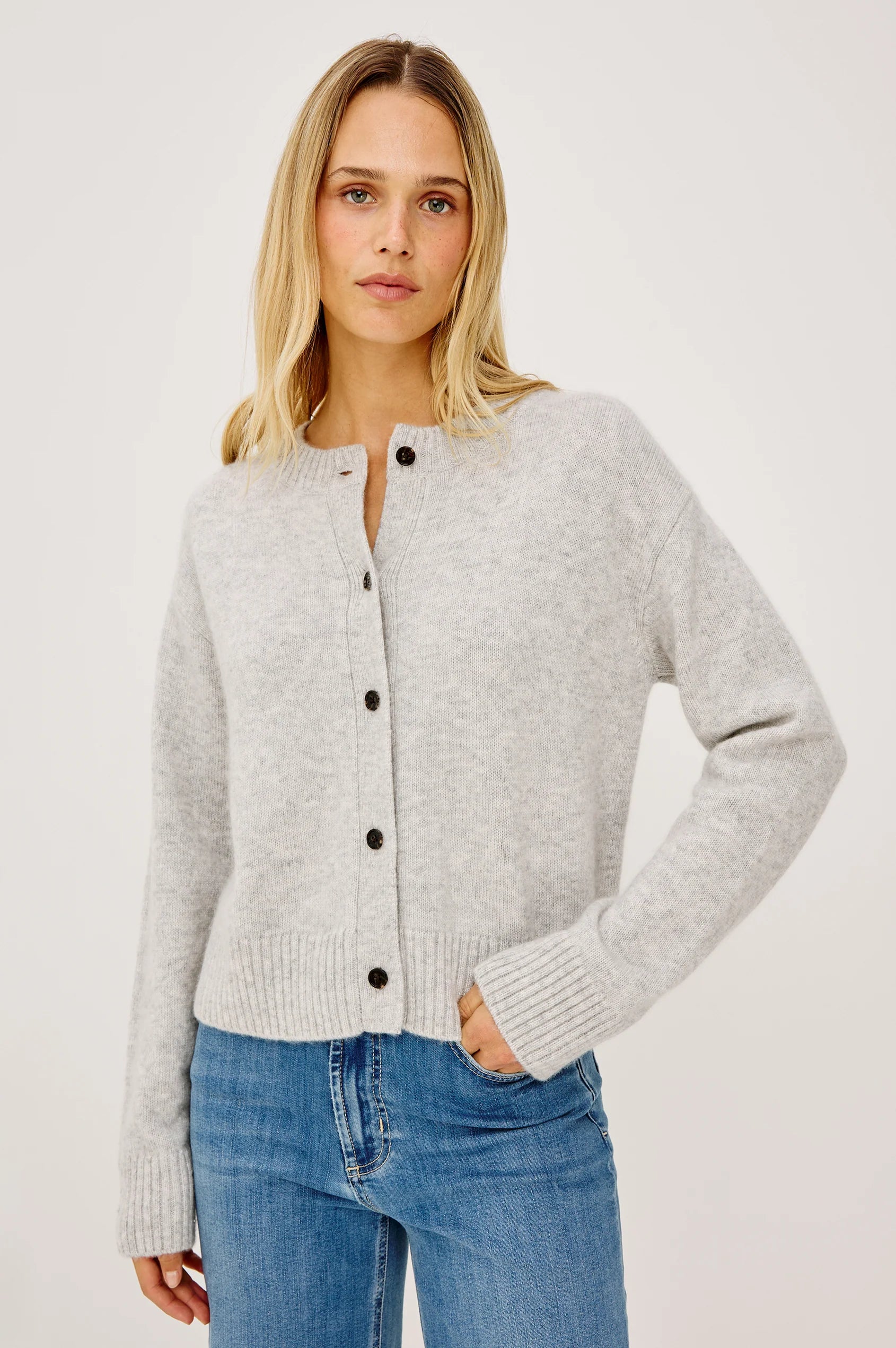 A woman wearing a light gray heathered knit cardigan with black buttons and blue jeans, photographed against a clean off-white studio background.