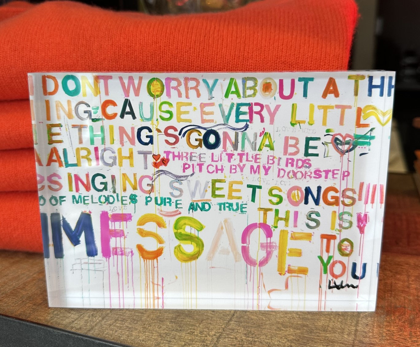 A clear acrylic block sign featuring colorful, hand-lettered text with a distressed paint-splatter background. The sign is resting on a dark wooden surface with a bright orange knit blanket behind it.