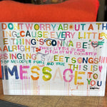 A clear acrylic block sign featuring colorful, hand-lettered text with a distressed paint-splatter background. The sign is resting on a dark wooden surface with a bright orange knit blanket behind it.