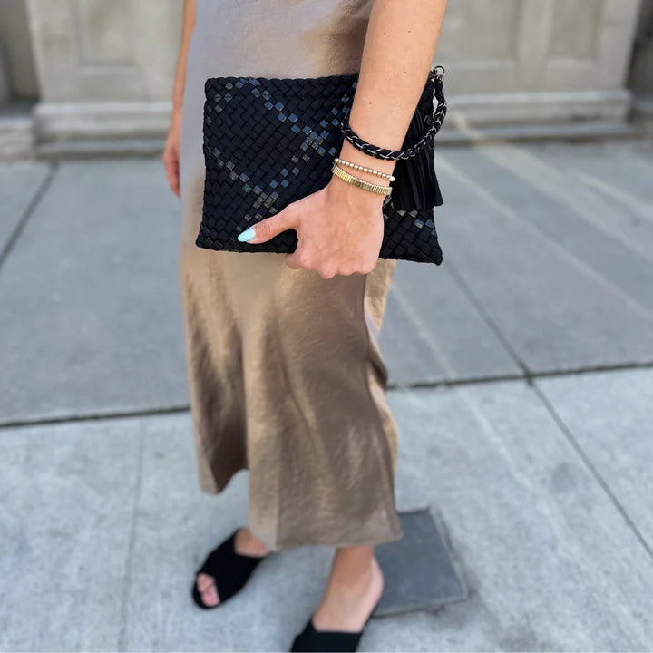 A person outdoors holding a black woven clutch bag with a braided chain strap. The person is wearing a taupe maxi dress, black slide sandals, and multiple bracelets.
