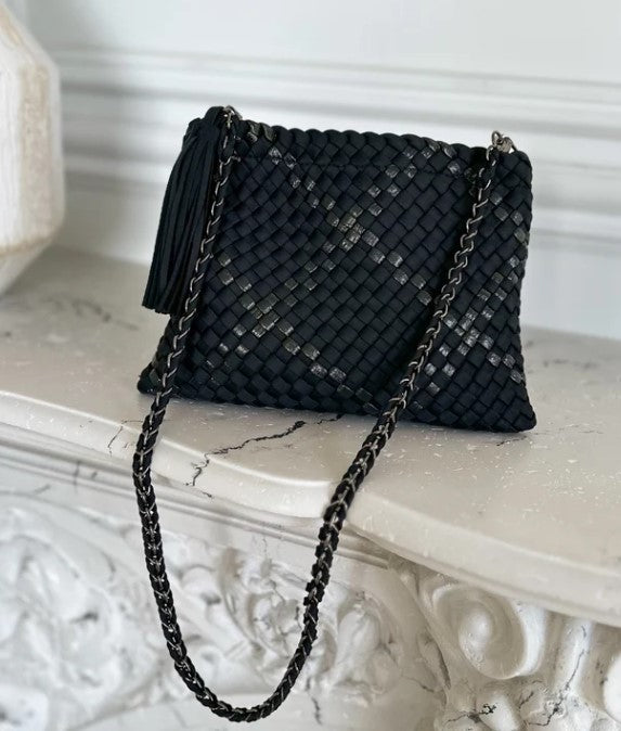 A black woven leather clutch bag with a tassel and a black-and-silver chain strap, resting on a white marble mantelpiece.