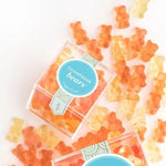 Orange and yellow gummy bears scattered on a white background with two clear plastic boxes of Champagne Bears gummies, featuring a blue circular label.
