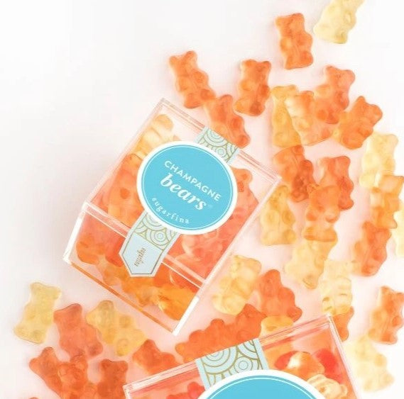 Orange and yellow gummy bears scattered on a white background with two clear plastic boxes of Champagne Bears gummies, featuring a blue circular label.