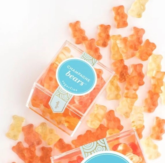 Orange and yellow gummy bears scattered on a white background with two clear plastic boxes of Champagne Bears gummies, featuring a blue circular label.